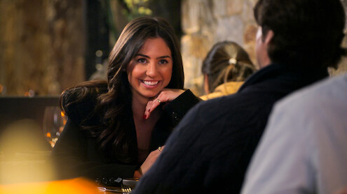 A woman smiles at a man across the table during a dinner in a warmly lit restaurant with stone walls and blurred background diners, creating a cozy and intimate atmosphere.
