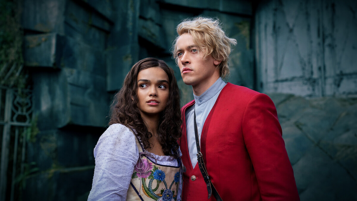 Two young adults in period costumes, a woman in a white dress with floral accents and a man in a red jacket, stand close together looking ahead, with a stone wall or ruins as the background, in a dramatic, moody setting.