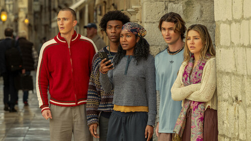 Five young adults stand together against a stone wall on a city street, looking ahead with focused and concerned expressions. The setting appears to be urban and historic, with wet pavement suggesting recent rain.