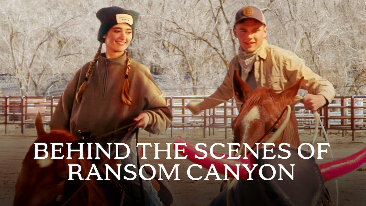 Da esquerda para a direita, Lizzy Greene e Garrett Wareing sorriem enquanto montam a cavalo em uma área cercada com árvores sem folhas. O texto na parte inferior diz “BASTIDORES DE RANSOM CANYON”.