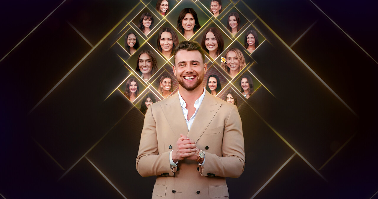 Um homem sorridente de terno bege está diante de um fundo escuro com losangos iluminados que exibem os rostos de várias mulheres acima e atrás dele, sugerindo o cenário de um reality show de namoro.