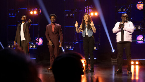 Four people stand on a stage with bright lights and spotlights behind them, in a lively performance or show setting, with one person pointing, two others smiling, and the group facing an audience.