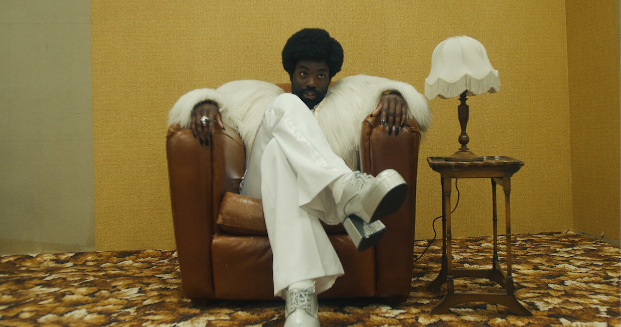 Janie Booth as Sweet Gran sits in a department store while Paapa Essiedu as Gaap wears a white fur coat standing behind her in Seaon 6 of Black Mirror.