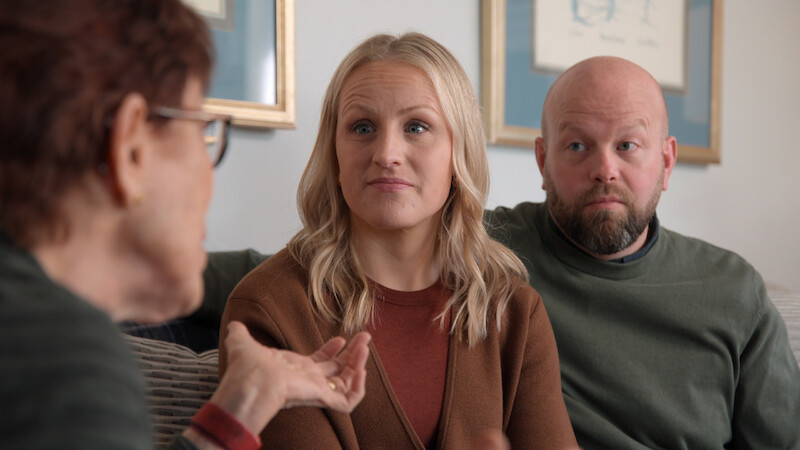 Three adults have a serious conversation while sitting on a couch in a living room, with framed artwork and soft lighting in the background.