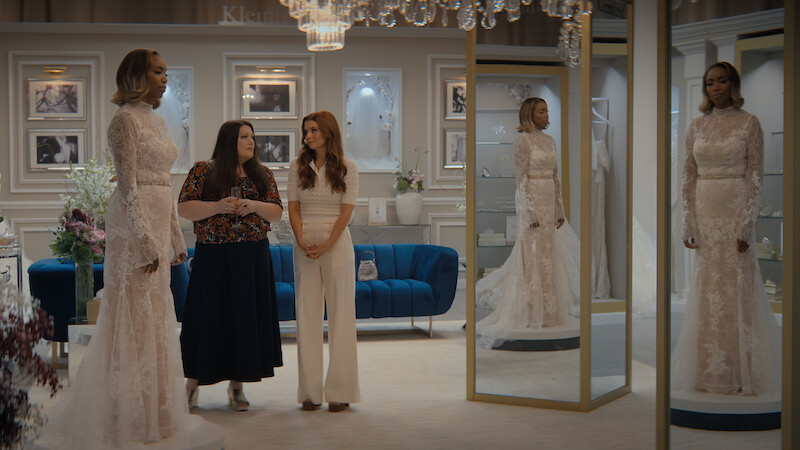 Three women in a stylish bridal shop, one wearing a wedding gown standing by mirrors, with elegant decor, framed photos on the wall, a blue couch, and chandeliers creating a classy atmosphere.