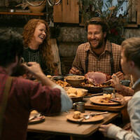 Four people share a rustic meal in a cozy, wooden cabin. The table is set with hearty food, and everyone is smiling and laughing, suggesting a warm, joyful gathering in a homey, inviting environment.