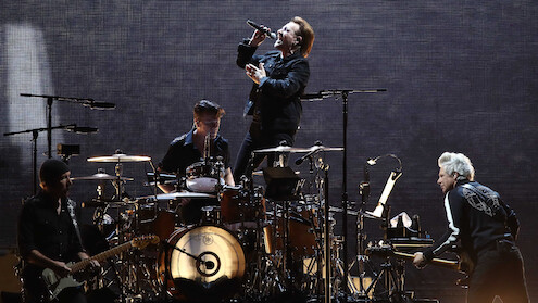 A rock band performs on stage with dramatic lighting; the lead singer stands on the drum riser singing into a microphone as other band members play guitar, drums, and keyboard in a concert setting.