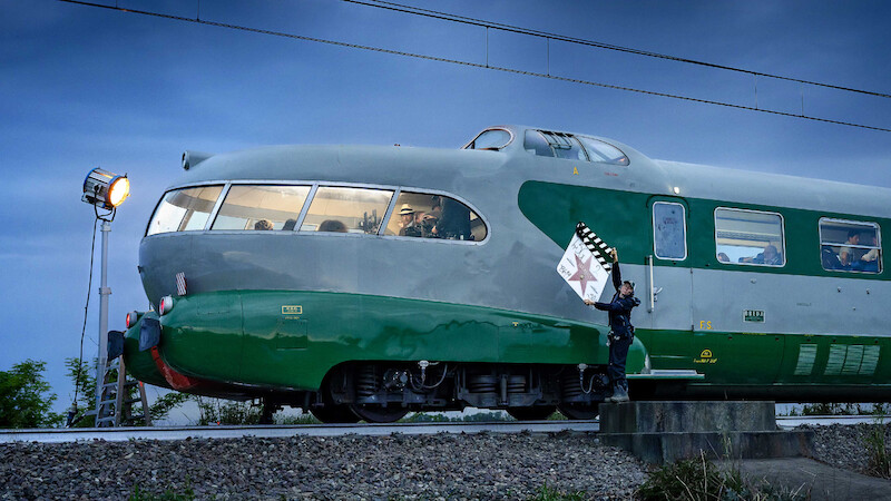 A vintage green and gray streamlined train is stopped outdoors at dusk on train tracks; a person in uniform stands beside it holding a clapperboard while a film light illuminates the scene, creating a cinematic atmosphere.