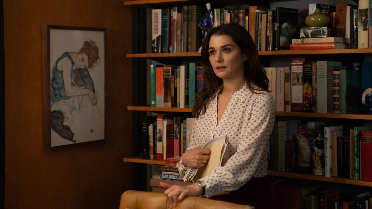 Rachel Weisz as The Protagonist  in a white blouse stands in front of a bookshelf filled with books and decor, holding folders. A framed artwork hangs on the wall, creating a cozy, intellectual office or library setting.