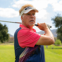 Man in a pink and blue polo shirt and visor holding a golf club in a follow-through pose on a sunny golf course with green grass, trees, and hills in background.