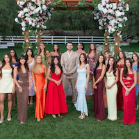 A group of elegantly dressed women and one man stand together outdoors on grass, beneath a floral archway, with trees and a white fence in the background, posing for a formal photo.