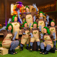 Grupo de pessoas em fantasias de umpa-lumpas, com perucas verdes, camisas marrons, leggings listradas e macacões castanho-claros posando e acenando no set de uma fábrica de doces colorida com guloseimas gigantes e maquinário extravagante ao fundo.