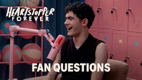 Joe Locke smiles and talks into a pink microphone in a colorful room with lockers and pastel basketballs, with "Heartstopper Forever" and "Fan Questions" text on screen, suggesting a fan Q&A interview setting.