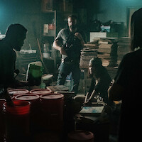 Armed officer and several people in a dimly lit, cluttered room with stacks of paper, buckets, and scattered objects, suggesting a tense or investigative scene in an industrial or basement setting.