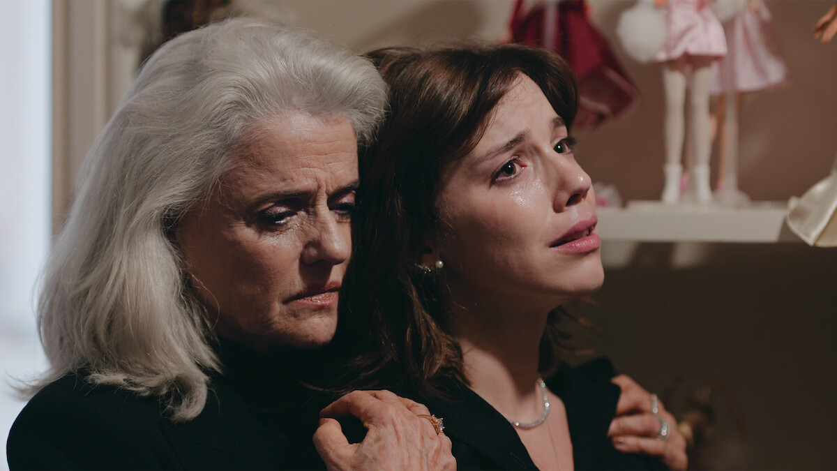As atrizes Marieta Severo, uma mulher branca de cabelos grisalhos, e Alice Wegmann, de cabelos curtos e castanhos, se abraçam num quarto com iluminação baixa e bonecas ao fundo