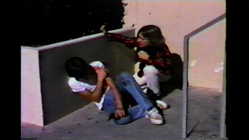 Charlie Sheen and Chris Penn are in a corner outside; one is sitting on the ground with their arm across their chest, while the other crouches nearby, reaching out toward them.