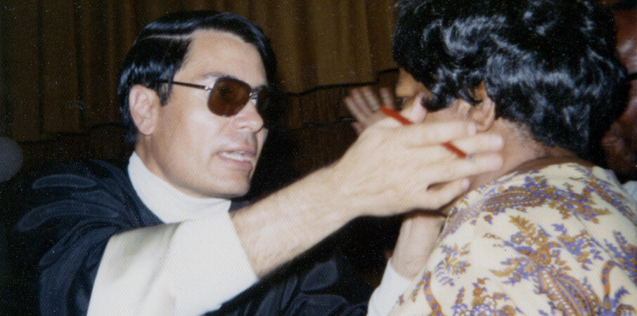 Man in glasses and dark robe extends his hands toward another person in a patterned shirt, indoors with brown curtain backdrop, suggesting an intense or dramatic interaction in a formal or ceremonial setting.