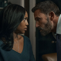 A serious man with a beard leans in closely toward a concerned woman in an indoor setting, both gazing intensely at each other, suggesting a tense or dramatic conversation.