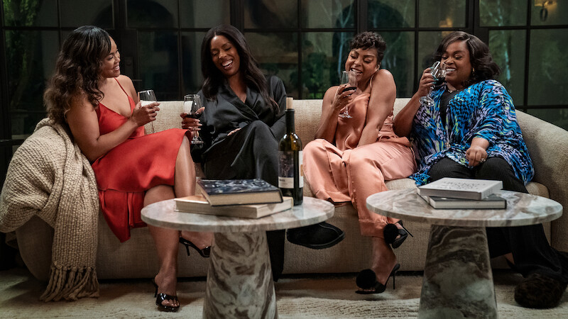 Four women sit on a sofa in a cozy living room, laughing and enjoying drinks together, with books and wine on marble tables in front of them, and large windows in the background.