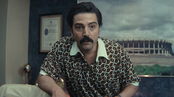 Man with dark hair and mustache in patterned shirt leans forward in an office with framed certificate on wall and large stadium photo in the background.