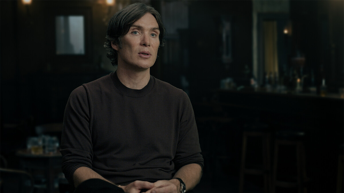 A man with short dark hair sits and speaks in a dimly lit bar or pub setting, with tables, chairs, and a bar counter visible in the background. The atmosphere is moody and subdued.