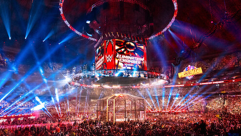 WWE Elimination Chamber wrestling event in a large arena, filled with a cheering crowd, colorful lights, a cage structure in the ring, and giant display screens with event logos and a WrestleMania sign in the background.