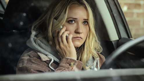 A young woman with blonde hair sits in a car, looking concerned while talking on a cellphone. She is wearing a hoodie and is positioned near the driver's seat, with a brick wall visible outside the window.