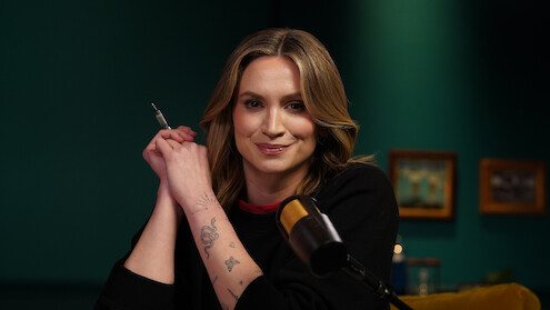 A woman with blonde hair and tattoos on her arm sits at a table in a studio, holding a pen, with a microphone in front of her. The background is teal with framed pictures on the wall.