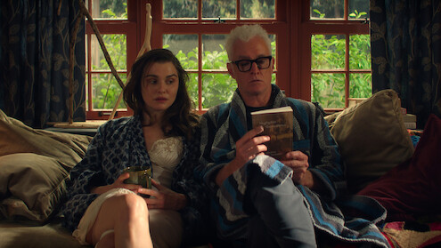 Rachel Weisz as M and John Slattery as John in bathrobes sit on a cozy couch by large windows, one holding a drink, the other reading a book, surrounded by cushions and natural light, with greenery visible outside.