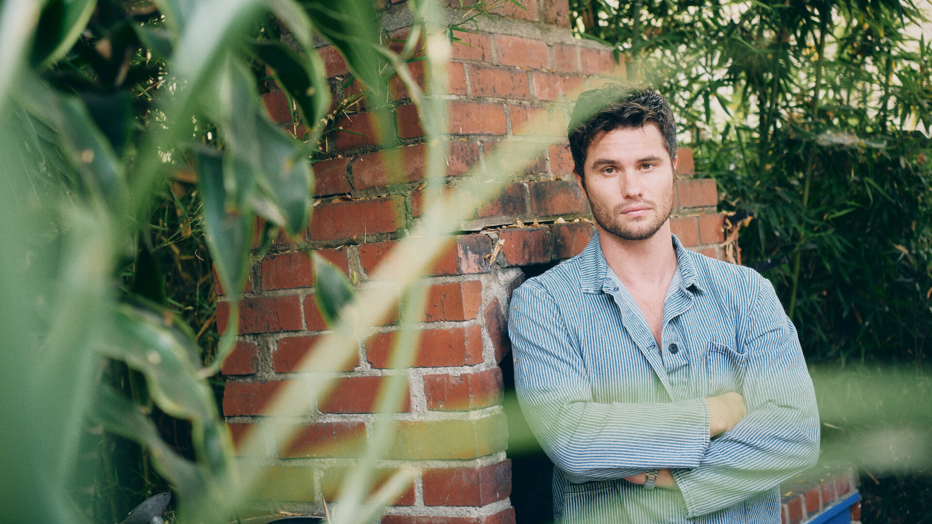 A man in a light denim shirt stands with arms crossed in front of a brick wall outdoors, surrounded by green plants and bamboo. The setting is natural and relaxed, with leaves partially obscuring the foreground.