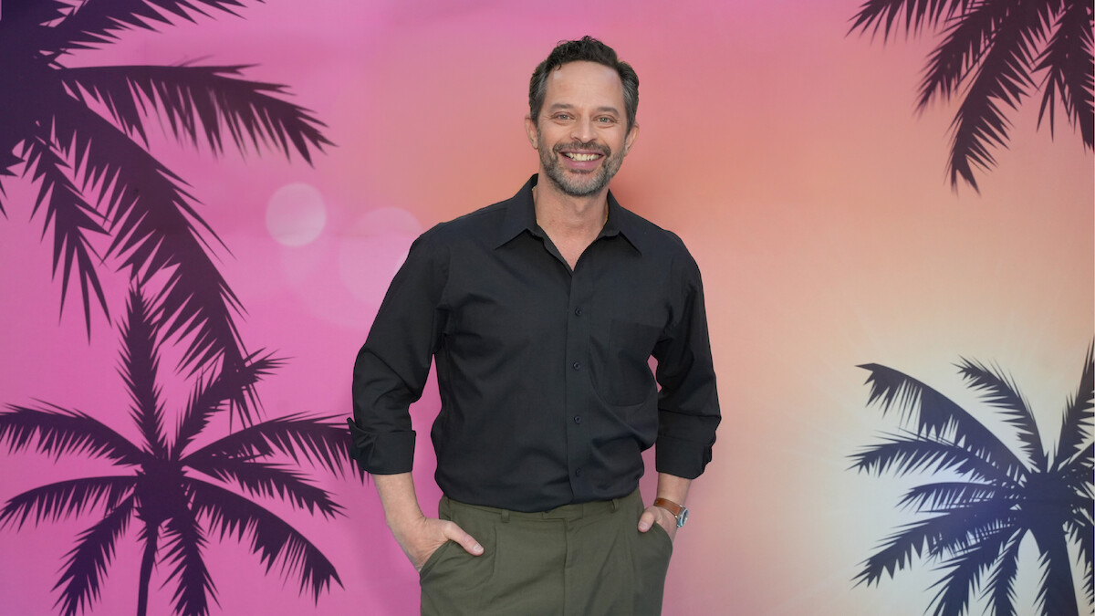Um homem sorridente, de camisa preta e calça verde, diante de um cenário de pôr do sol colorido, com silhuetas de palmeiras, evocando um ambiente tropical ou de praia.