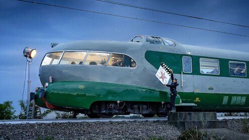 A vintage green and gray streamlined train is stopped outdoors at dusk on train tracks; a person in uniform stands beside it holding a clapperboard while a film light illuminates the scene, creating a cinematic atmosphere.