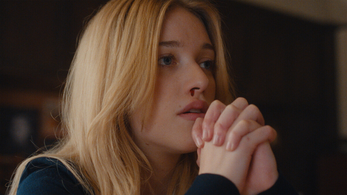 Young woman with long blonde hair sitting indoors, resting her chin on clasped hands, looking thoughtful or concerned, with a blurred background and warm lighting.