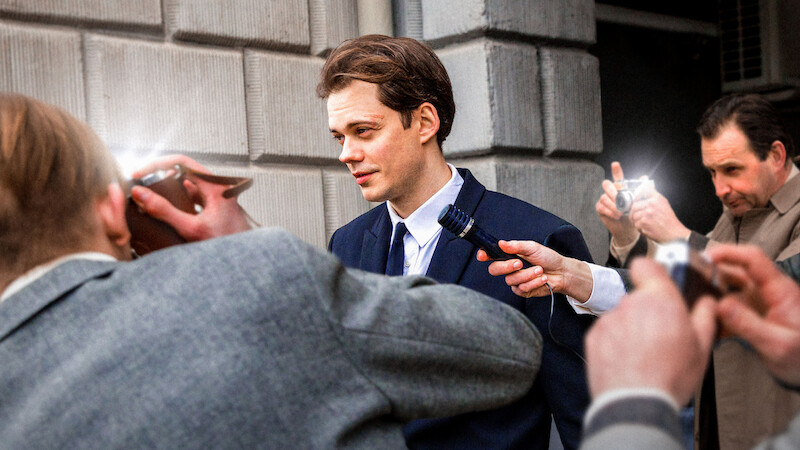 Bill Skarsgård in a suit as a character facing the press outside a grey brick building