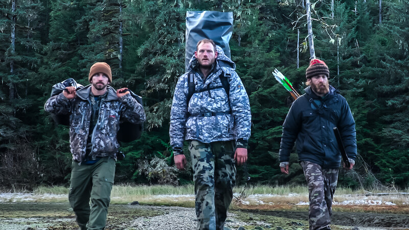 Three 'Outlast' Season 2 contestants walk towards the camera with their equipment and supplies.