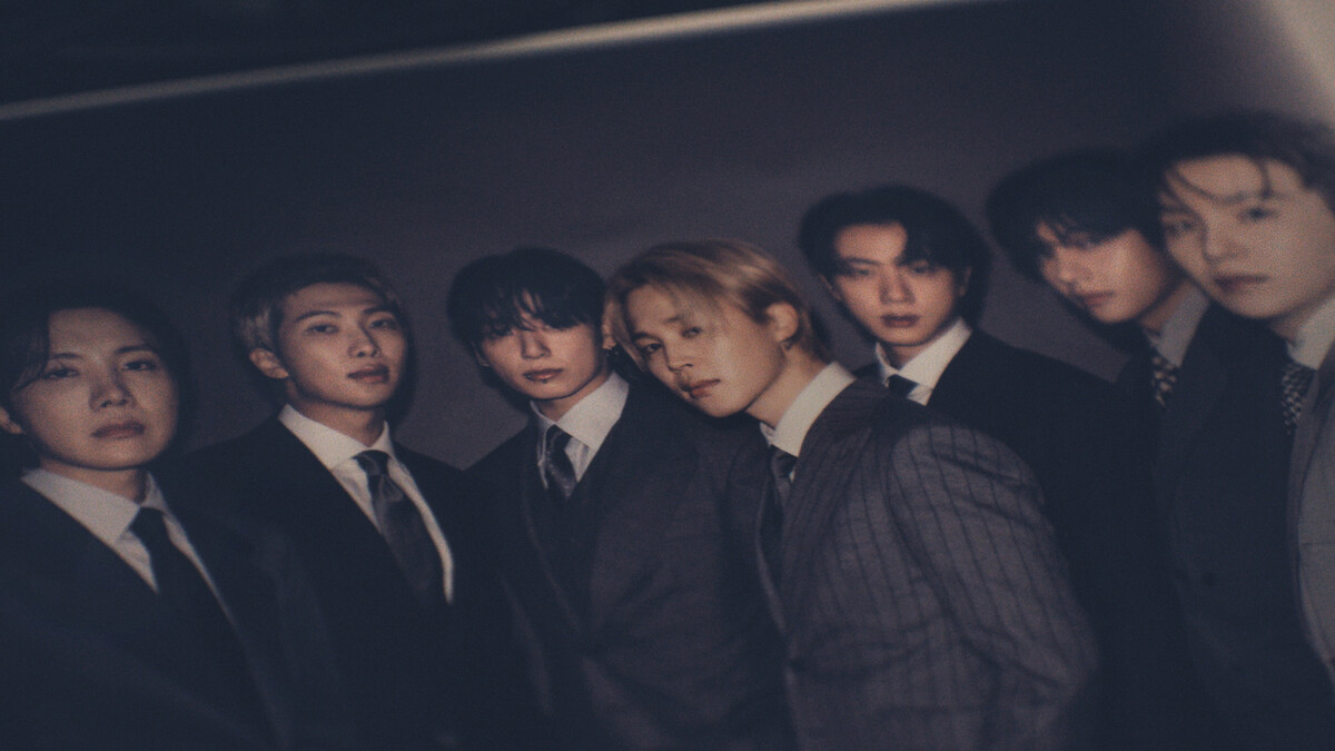 Seven young men wearing formal suits pose together in a dimly lit, dark environment, standing closely for a group portrait with serious and calm expressions.