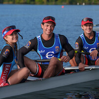 Three smiling rowers in team uniforms sit in a racing shell on a calm lake, wearing hats and sunglasses, with trees and water in the background, ready for a rowing competition or practice.