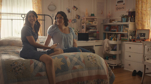 Two women sitting on a bed in a cozy, sunlit bedroom with bookshelves, photos, and personal items in the background, creating a warm and inviting atmosphere.