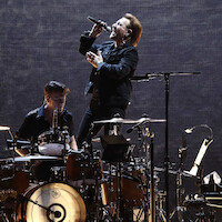 Lead singer passionately performing into a microphone on stage, with a drummer playing behind him, surrounded by musical instruments and equipment, under dramatic lighting against a dark background.