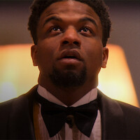 A man in a tuxedo and bowtie looks upward in a warmly lit indoor setting, appearing thoughtful or contemplative. The background is softly blurred, enhancing the formal and introspective mood of the scene.