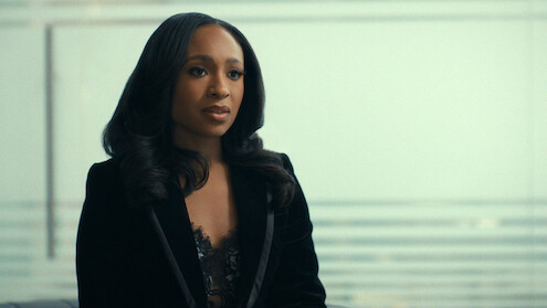 Woman with long dark hair in a black blazer sits indoors with a blurred glass wall and horizontal lines in the background, suggesting a modern office or professional setting.
