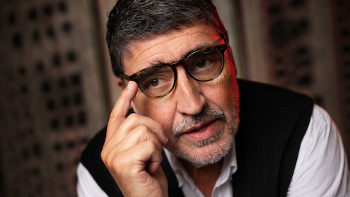 Alfred Molina with his hand raised to his glasses wearing a sweater vest and white button up shirt.