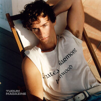 Young man with curly hair lounging on a deck chair indoors, wearing a white "Chateau Marmont Hollywood" t-shirt and jeans, sunlight highlighting his face and casting shadows on the wooden floor.