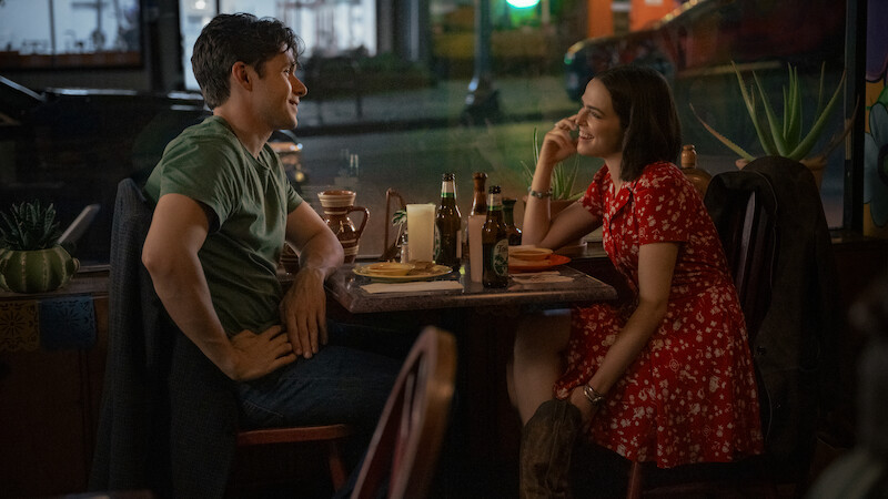 A man and woman sit across from each other at a cozy restaurant table at night, smiling and talking over food and drinks, with a street visible through the window behind them.
