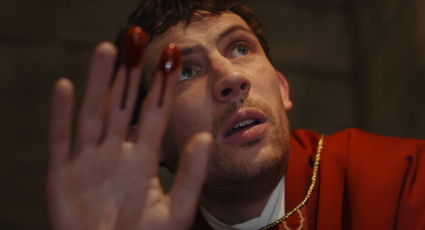 A worried man in a red robe holds up his hand with blood on his fingers, looking upwards in a dimly lit, stone-walled setting, creating a tense and dramatic mood.