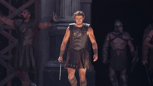 A group of male gladiators in ancient Roman armor stand in a dramatic, dimly-lit stone arena, with one warrior holding a sword at center stage, looking intense and determined.