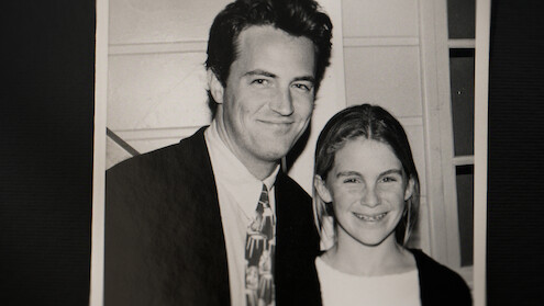 A black and white photo of a man in a suit and a young girl standing side by side, smiling, indoors with a paneled wall and window in the background.