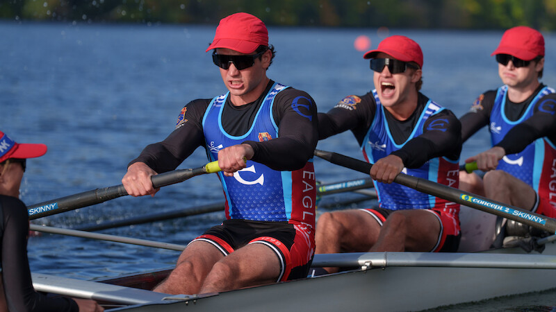 Rowers in matching uniforms and red hats compete in a crew race on a calm river, focused and working together in an outdoor setting with greenery in the background.