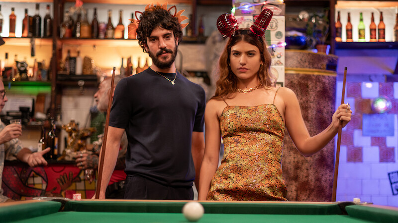 Um homem de camiseta preta, barba e cabelos grandes, e uma mulher de cabelos longos e castanhos, vestindo um vestido amarelo, se posicionam na frente de uma mesa de sinuca com a bola branca centralizada no primeiro plano