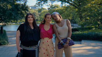 Three women standing close together outdoors on a sunny day in a lush park, smiling and posing for a photo with trees, greenery, and a body of water in the blurred background.